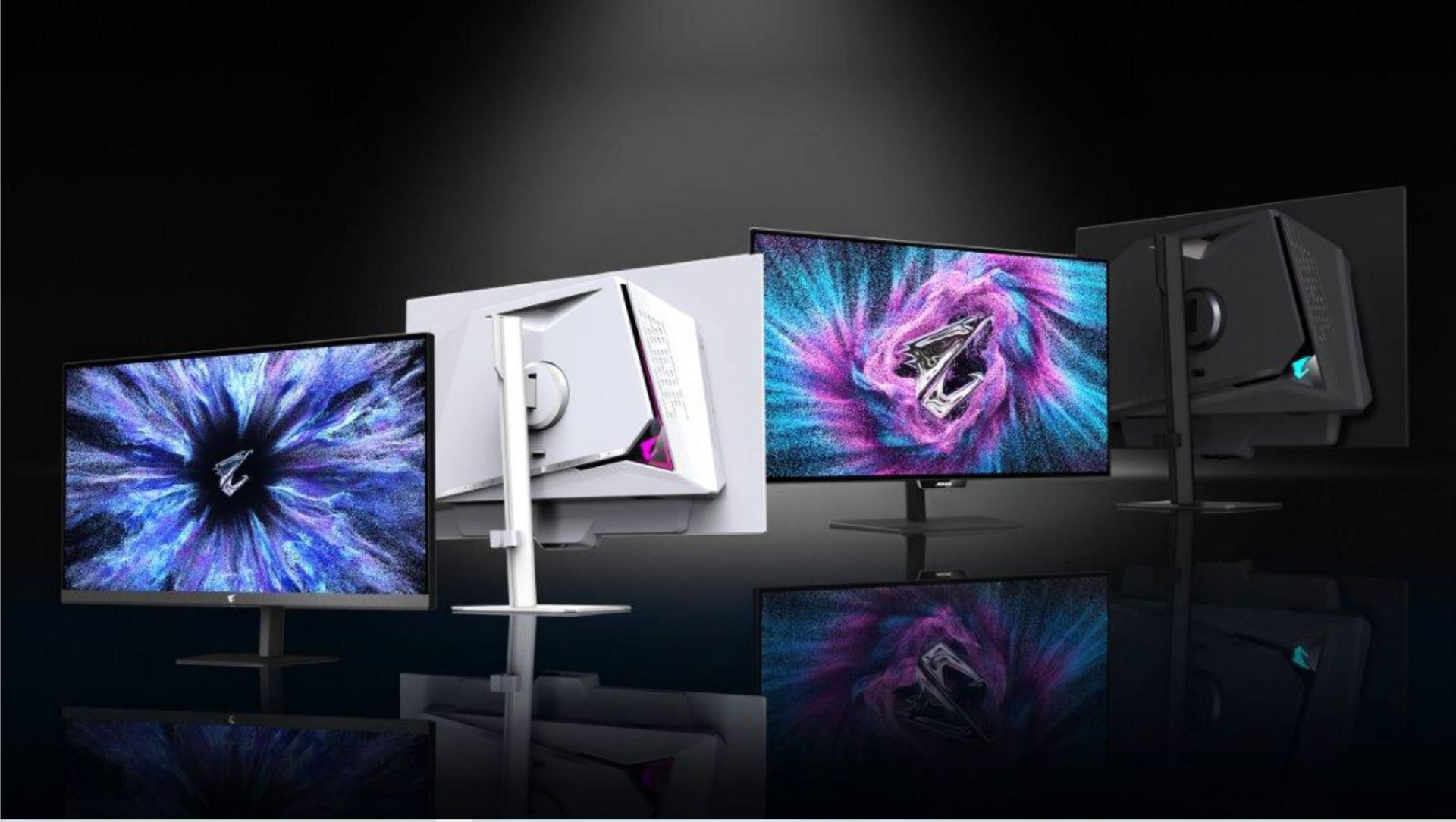 Three AORUS gaming monitors are displayed, two facing forward with vibrant abstract designs on the screens and one showing the side and back design, featuring angular stands and sleek back panels.
