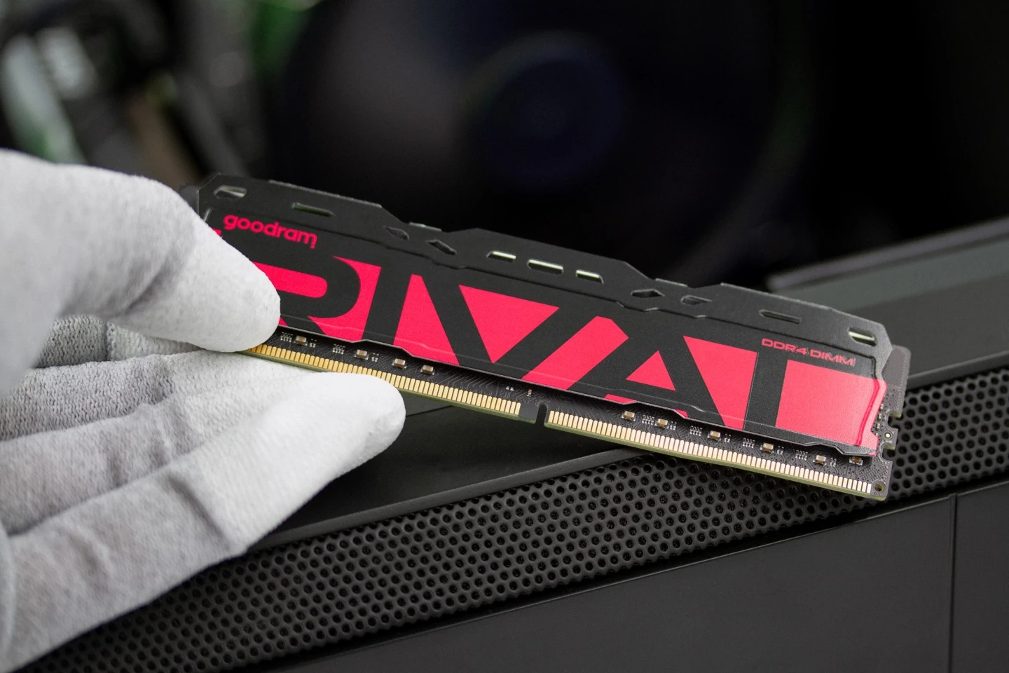 A person wearing gloves holds a Goodram DDR4 DIMM module labeled 'RIVAL' above a computer case.