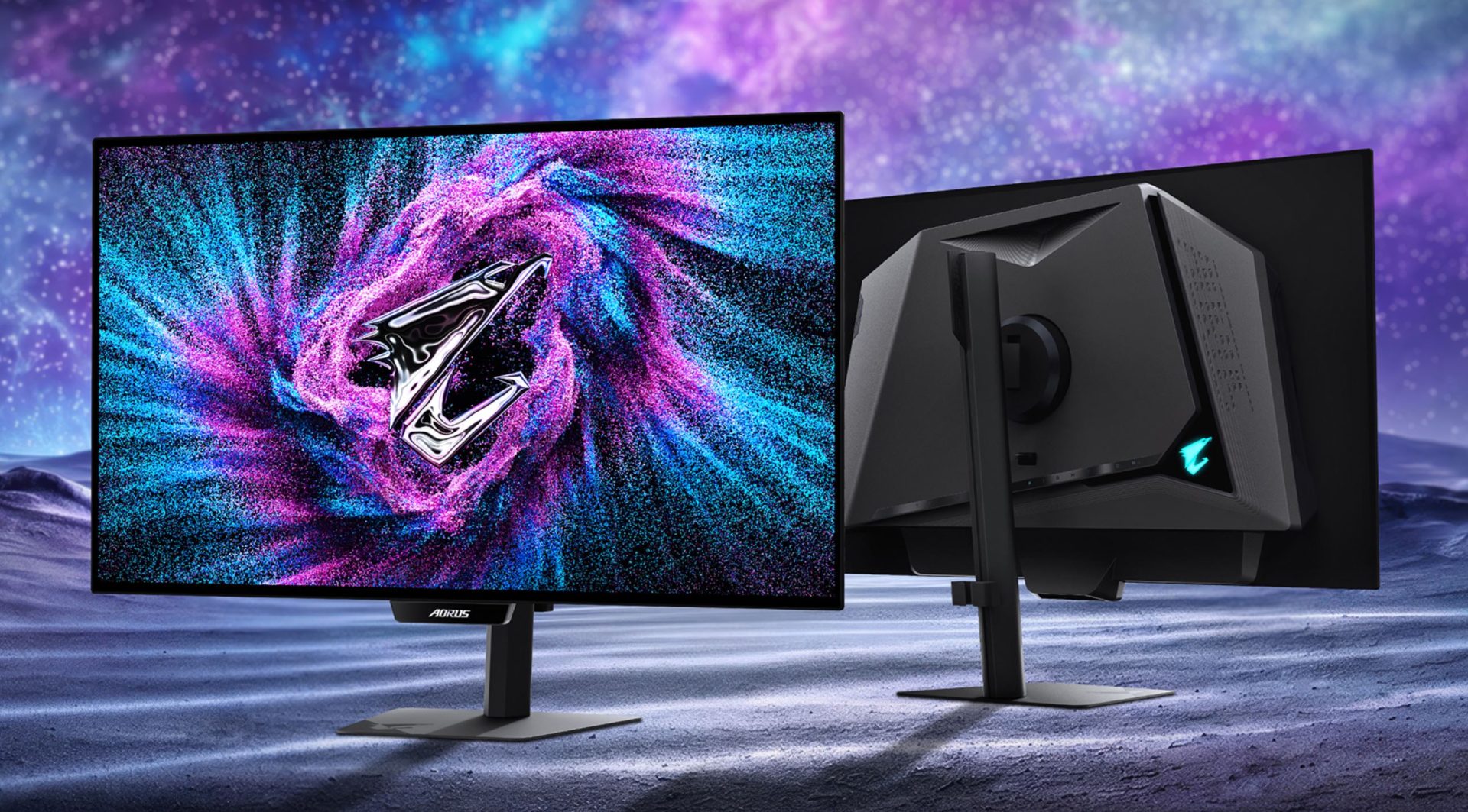 The image shows Gigabyte Aorus gaming monitors with a vibrant cosmic-themed display and the Aorus logo prominently featured on both the screen and monitor stand.
