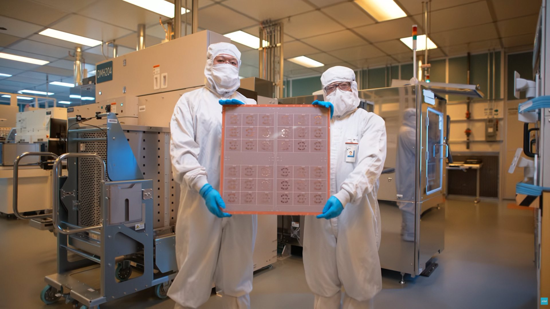 Two people in cleanroom suits holding a semiconductor wafer in a fabrication facility.