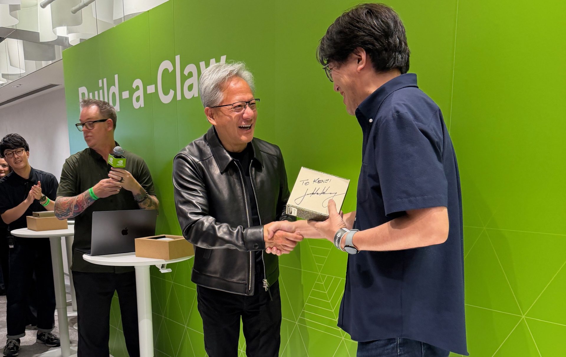 Two people shaking hands in front of a green wall with 'Build-a-Cluster,' as one holds a signed item inscribed 'To Keoni'.