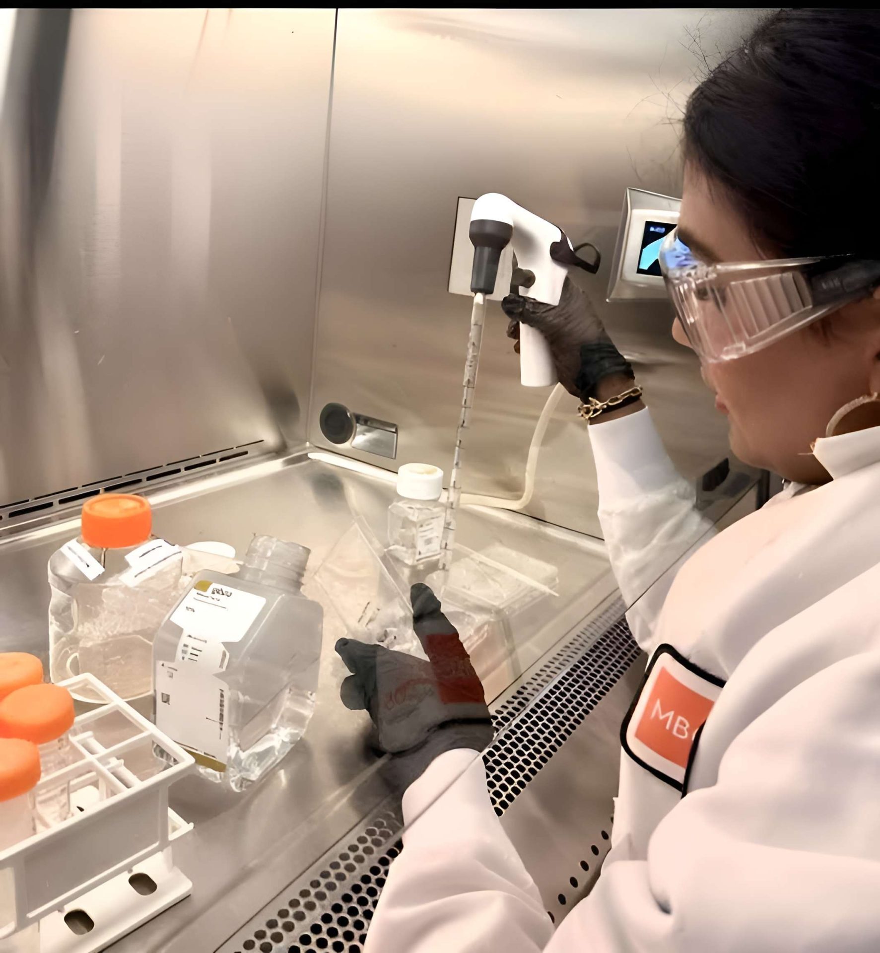 A lab technician wearing protective glasses and gloves uses a pipette inside a sterile cabinet, with MB visible on her lab coat, surrounded by containers filled with clear liquid.