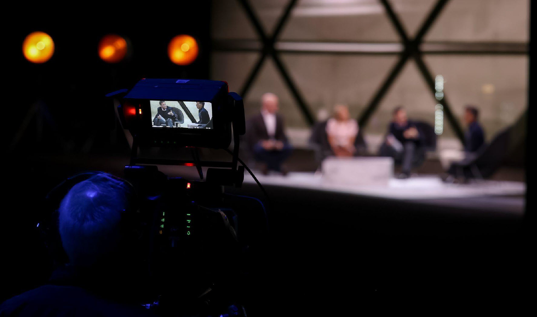 A broadcast camera captures a panel discussion at an event, with four blurred figures sitting on stage in the background.