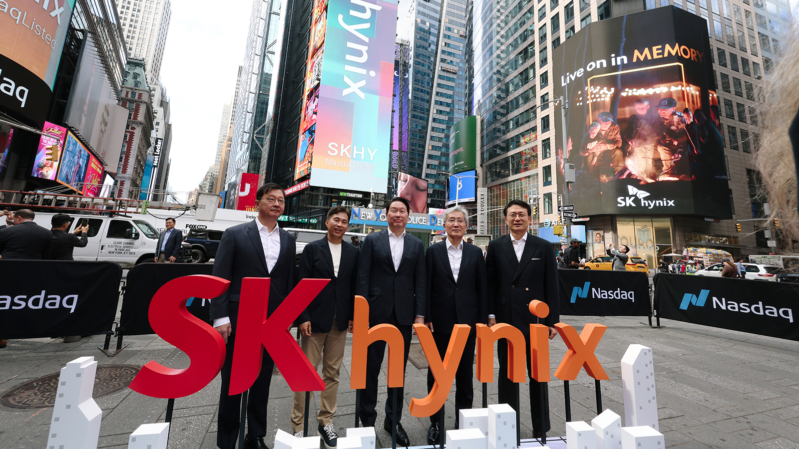A group of executives stands in front of SK hynix signage at Times Square, with a large screen displaying 'Live on in MEMORY' and the Nasdaq logo visible.
