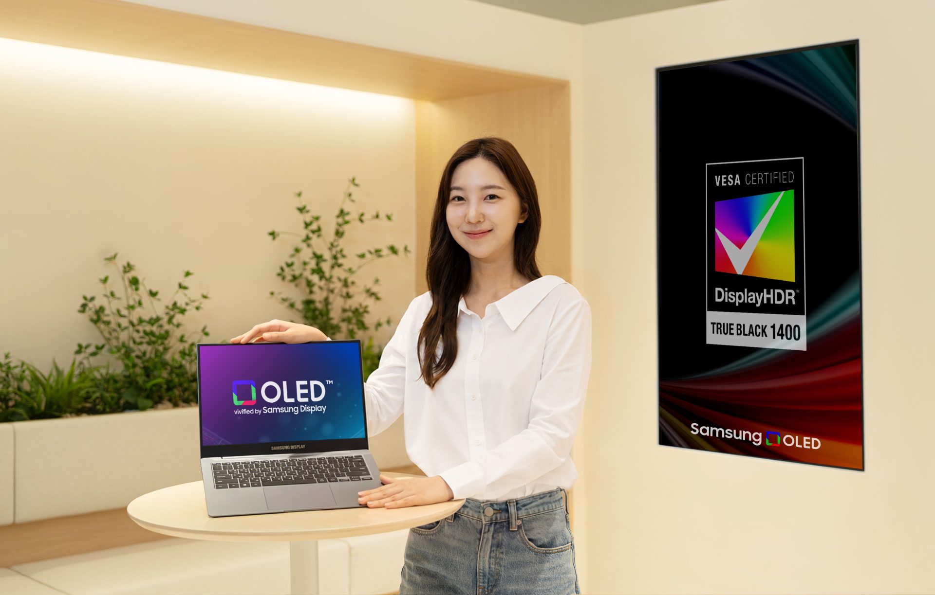 A person is holding a Samsung Display laptop featuring an 'OLED' screen in a room with a poster displaying 'VESA Certified DisplayHDR True Black 1400' and 'Samsung OLED' branding.