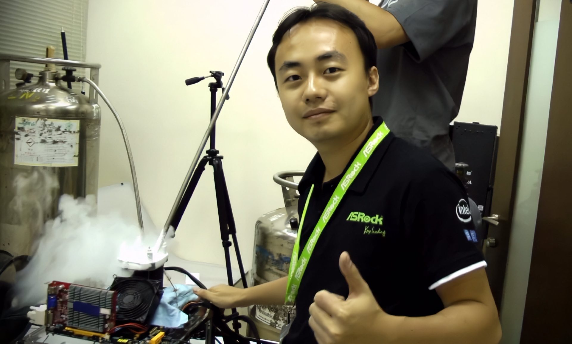 A person wearing an ASRock shirt gives a thumbs up next to a setup involving liquid nitrogen cooling equipment in a hardware testing environment.