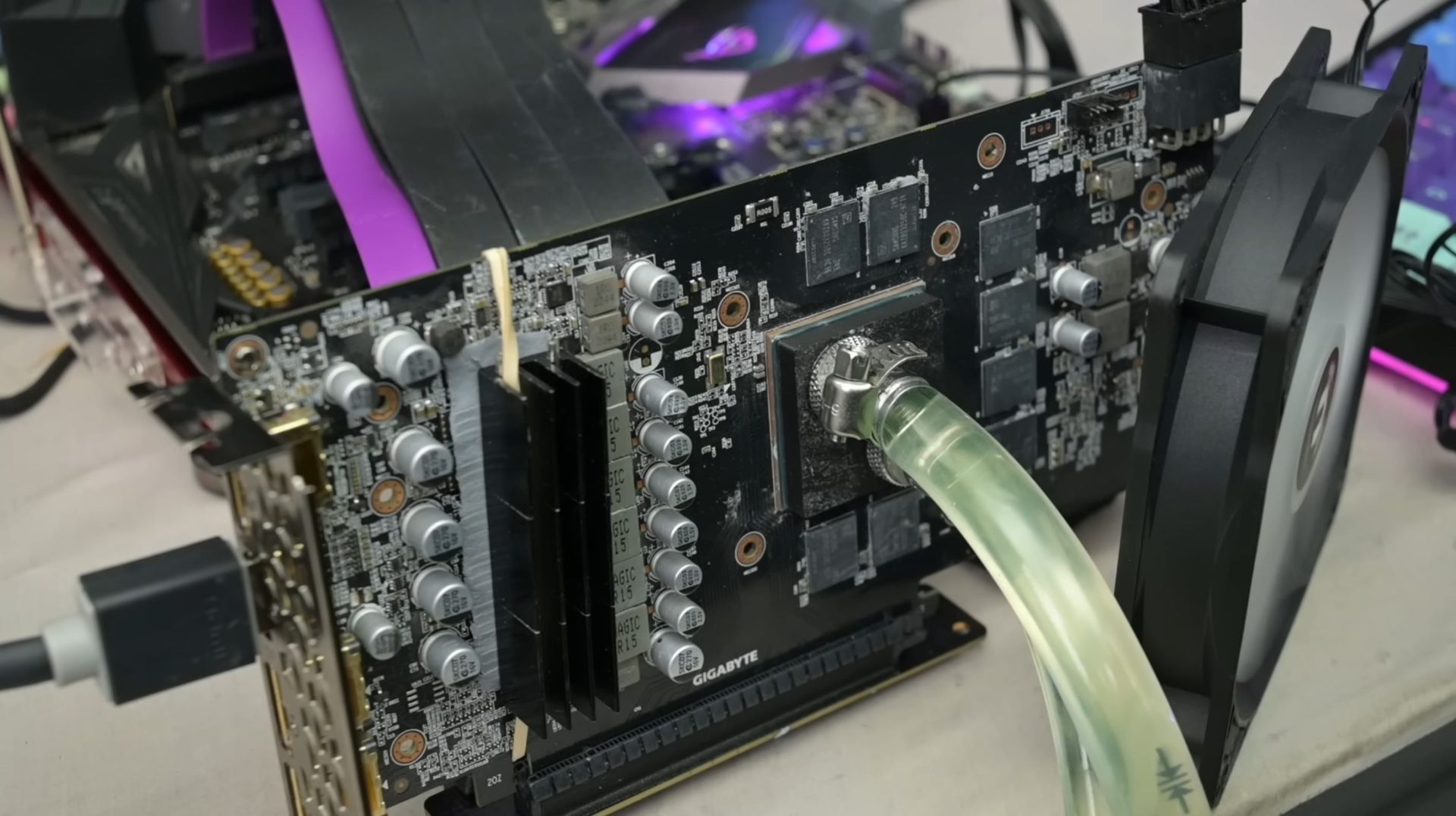 A Gigabyte graphics card with liquid cooling tubing is connected to a custom cooling setup, showing VRM components and power connectors on a test bench.