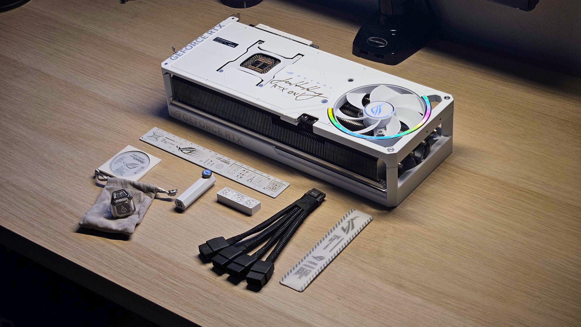 A custom white NVIDIA GeForce RTX graphics card with accessories, including cables and branding items, is displayed on a wooden desk.