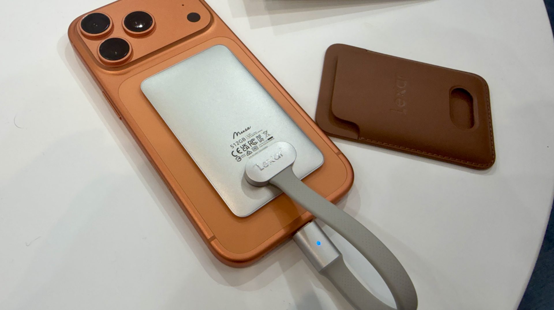 A Lexar portable SSD marked '512GB' is connected to an orange smartphone with a case nearby.