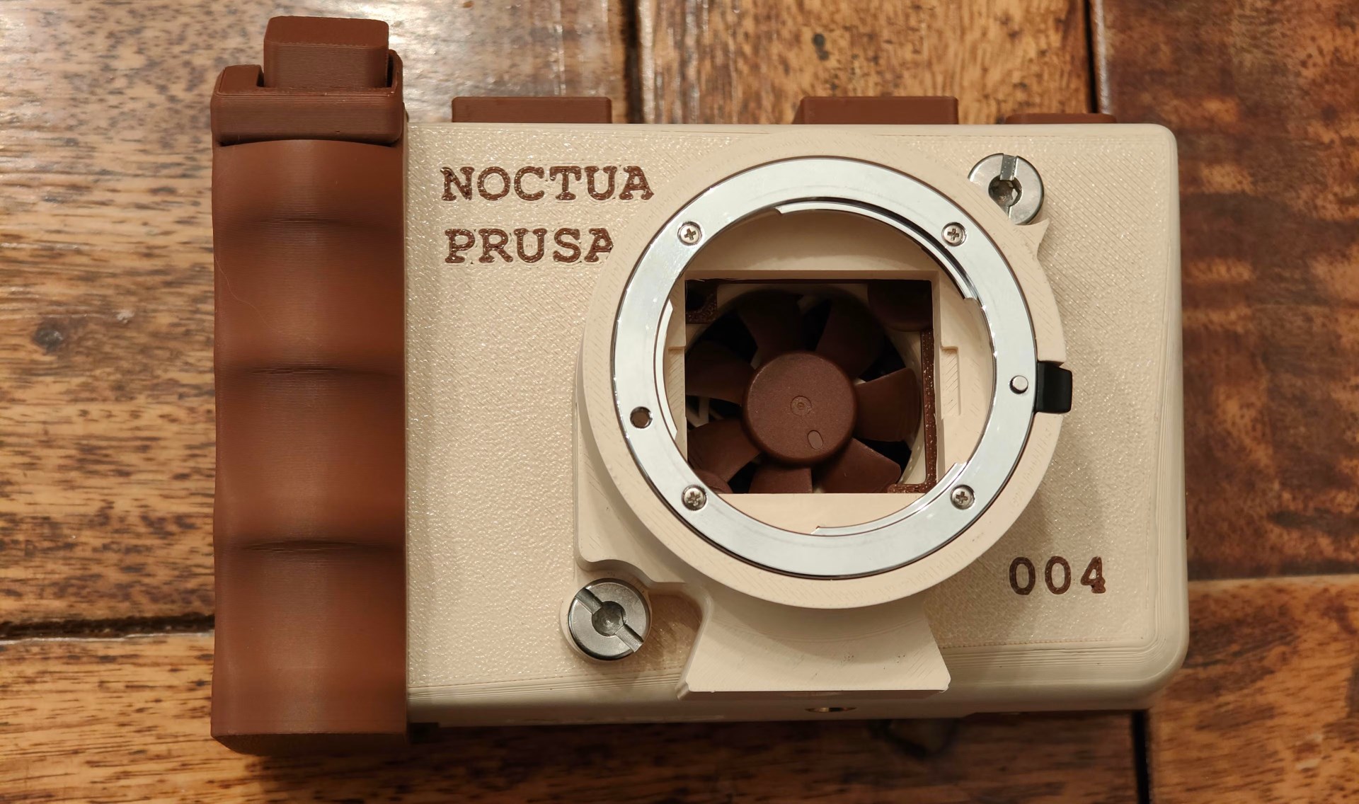 A custom camera case with a visible fan and 'NOCTUA PRUSA' text on a wooden surface, marked with '004'.