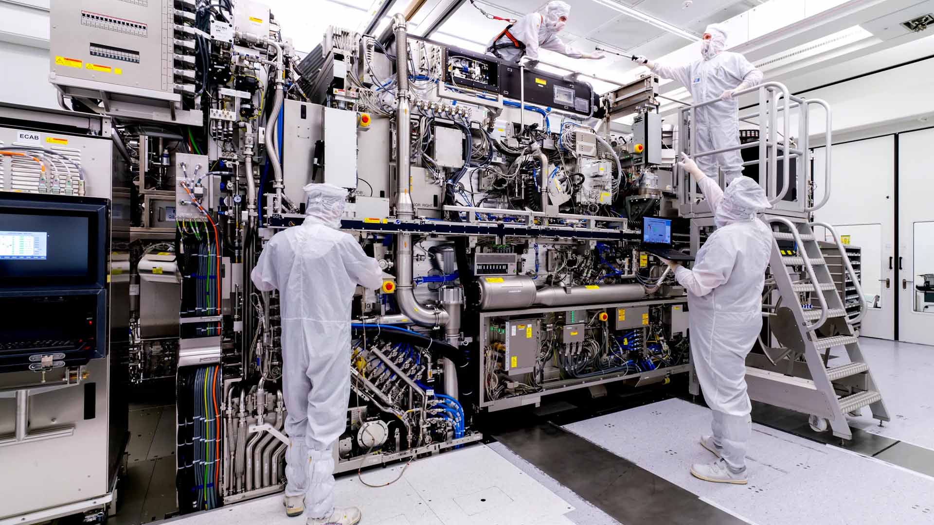 Technicians in cleanroom suits operate and examine a large, complex piece of semiconductor manufacturing equipment with visible cables, control panels, and monitors.