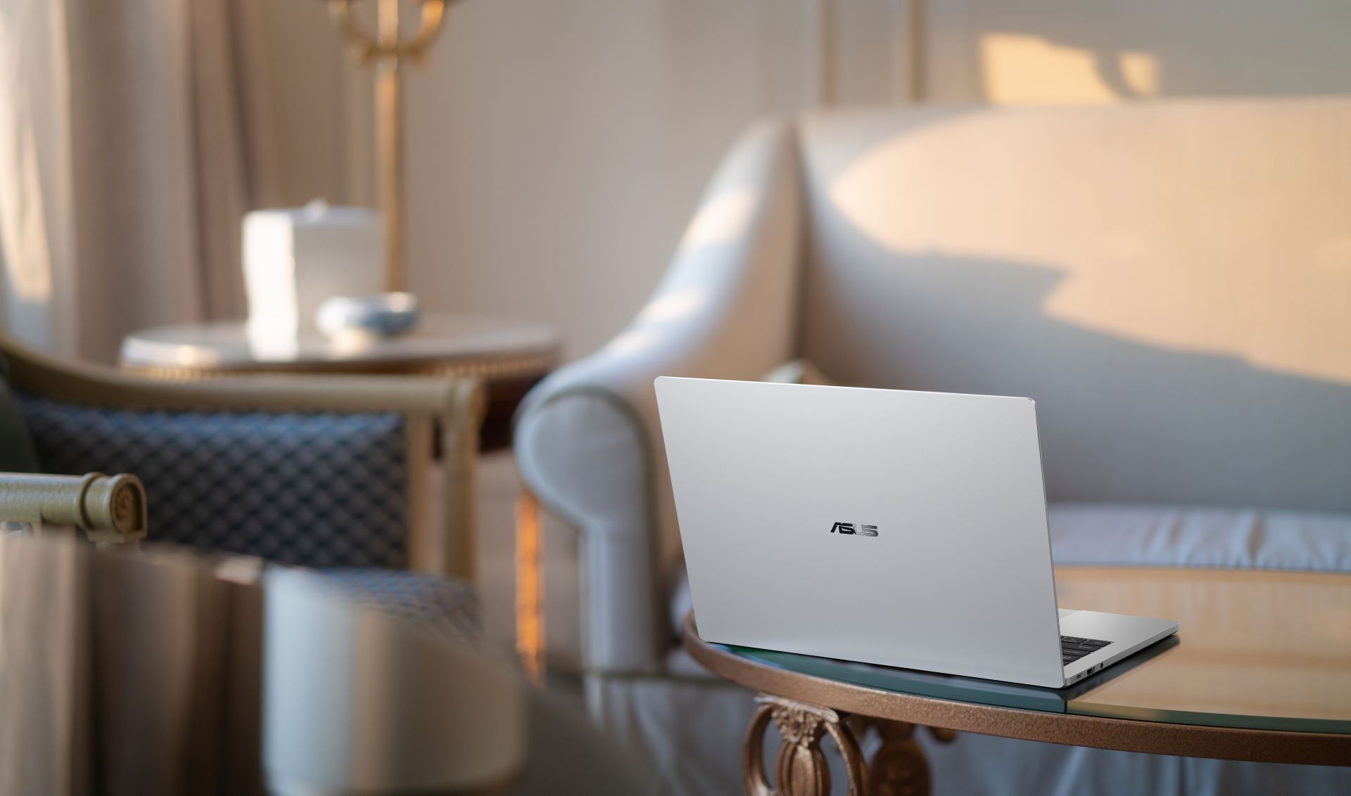 An ASUS laptop with a silver finish is placed on a glass table in a softly lit room with a sofa and round side table in the background.