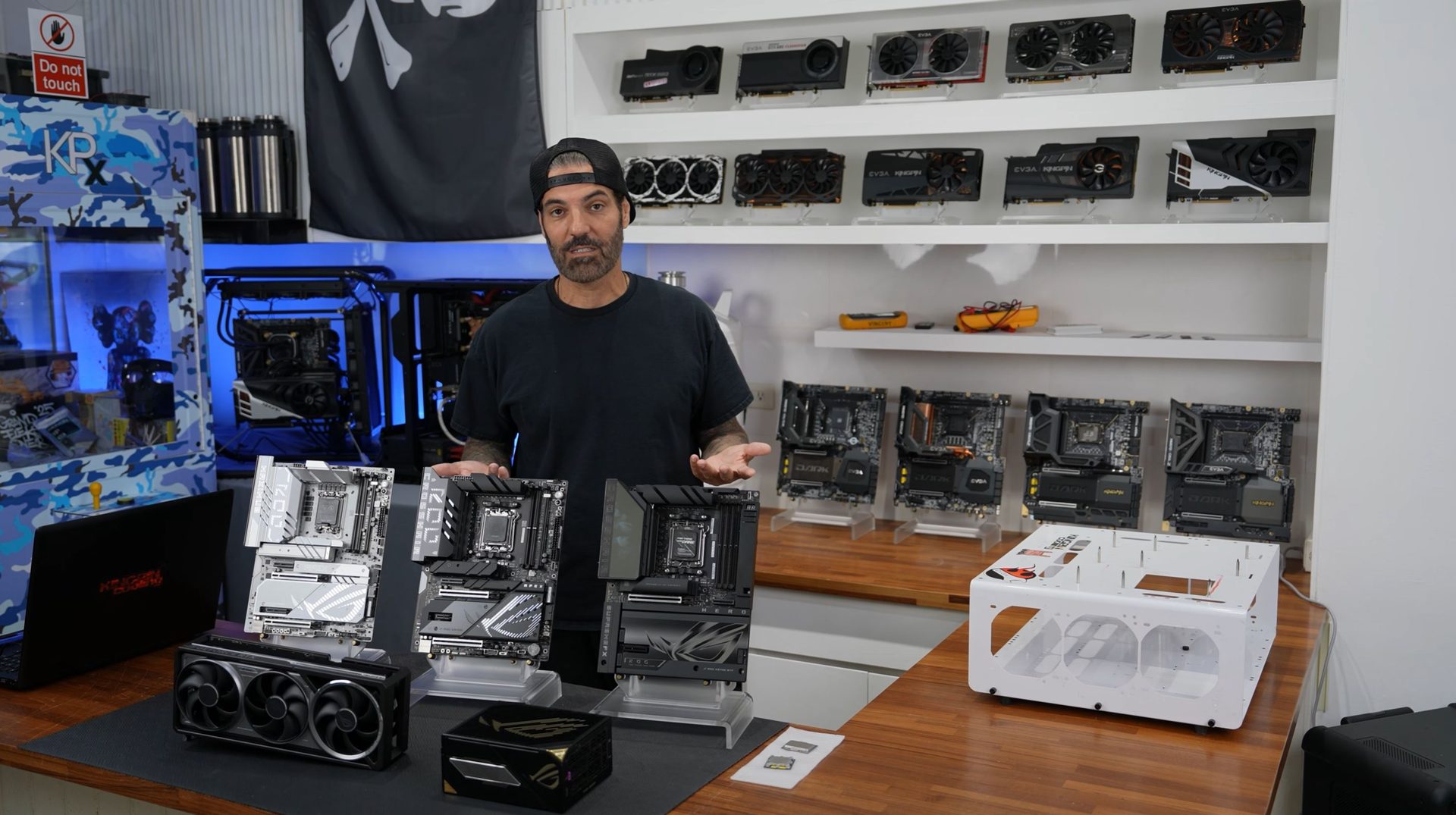 A person showcases several motherboards and a GPU on a table, with shelves displaying various graphics cards in the background.