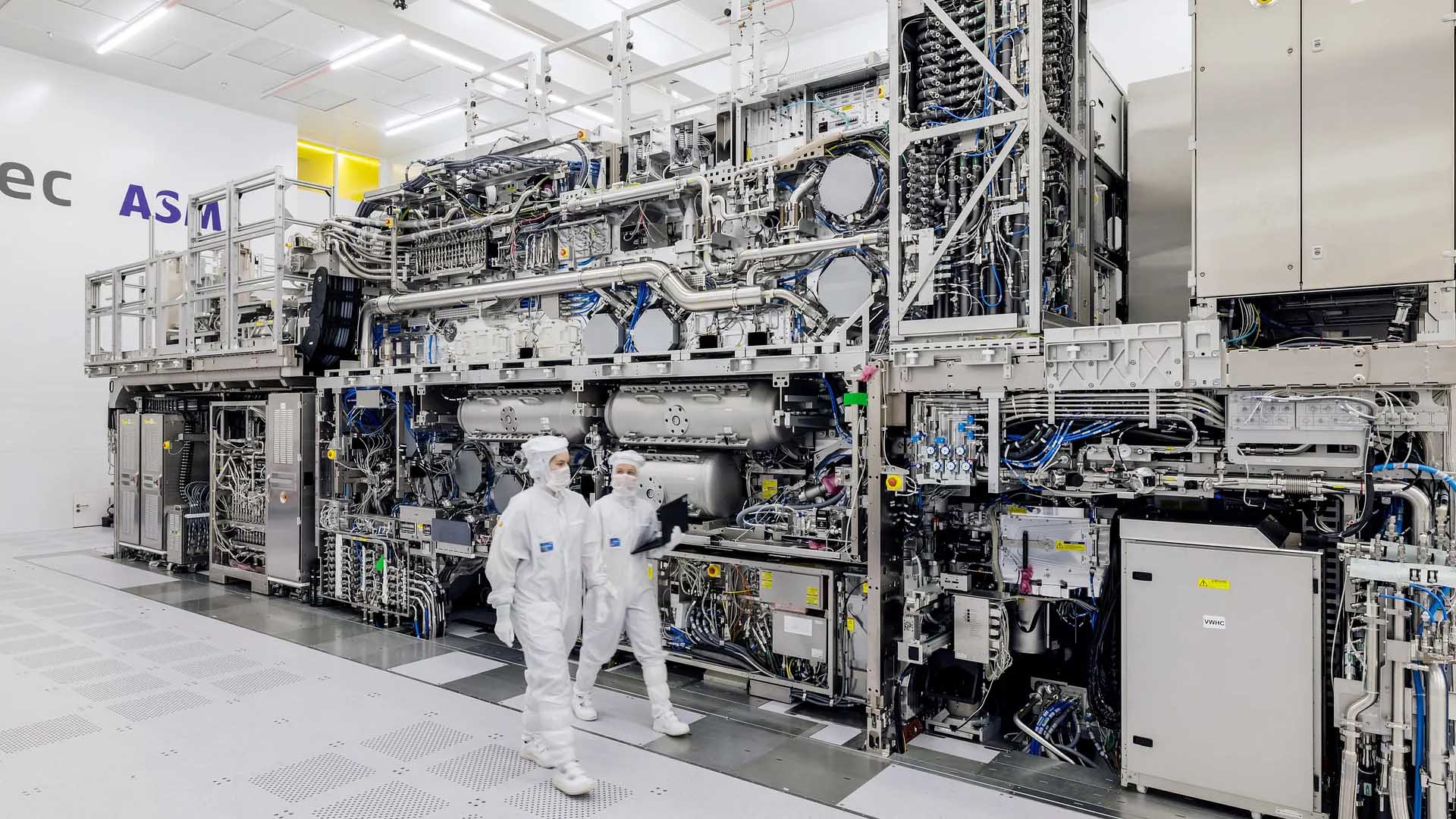 Two technicians in protective suits walk past a large ASML semiconductor manufacturing machine inside a clean room.
