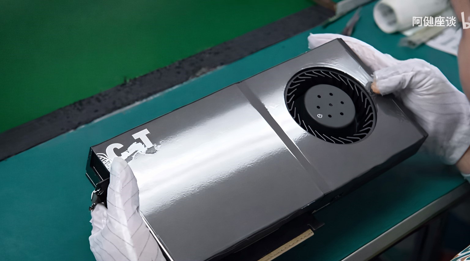 A person wearing gloves holds a graphics card labeled GT with a circular cooling fan on a green workbench.