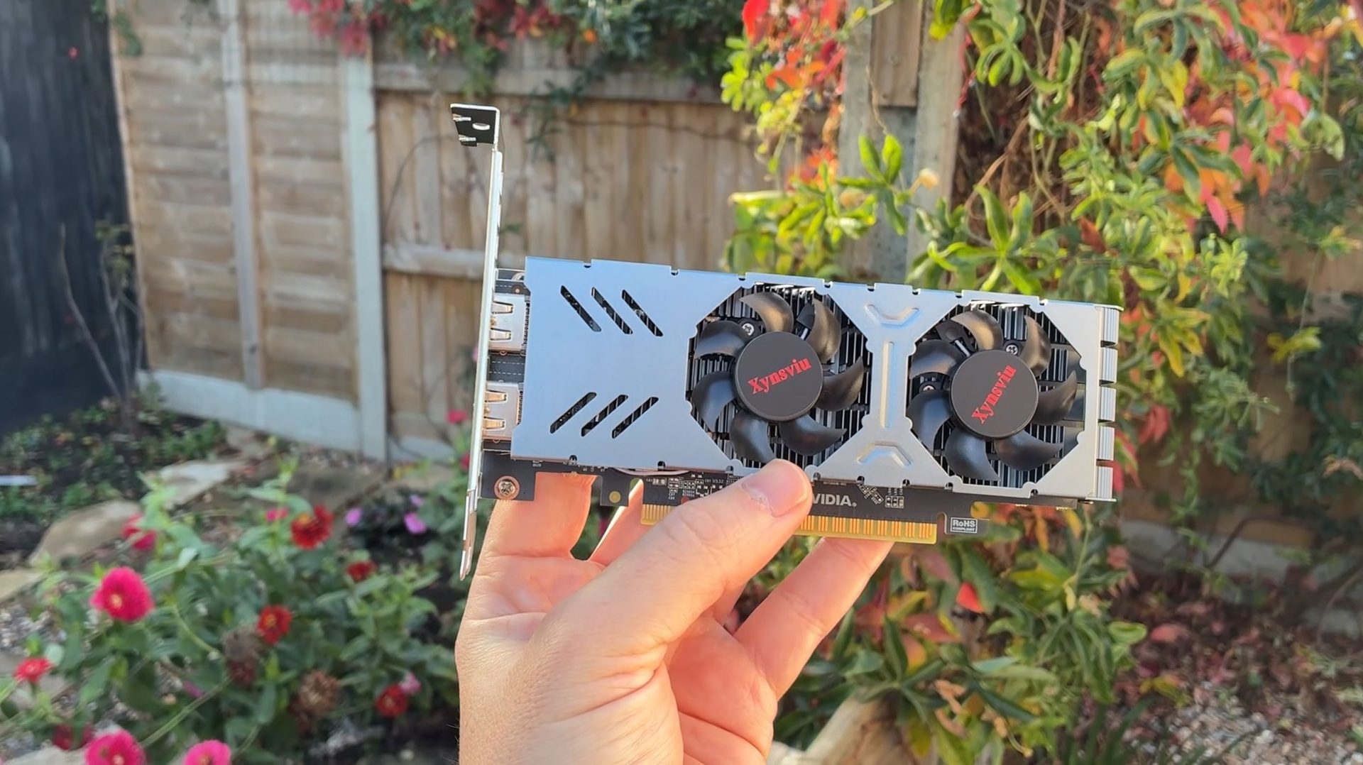 A hand holds a graphics card with a visible Nvidia logo, dual fans labeled 'Xynsviu,' and a PCIe interface, set against a garden backdrop.